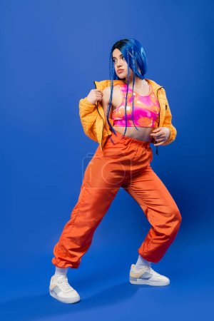 longitud completa, cabello teñido, moda y estilo, modelo femenino con pelo azul posando en chaqueta hinchable y pantalones naranja sobre fondo azul, color vibrante, moda urbana, individualismo, mujer joven 