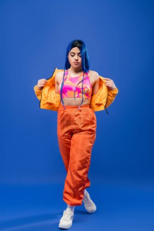 full length, dyed hair, modern style, female model with blue hair posing in puffer jacket and orange pants on blue background, vibrant color, urban fashion, individualism, young woman 