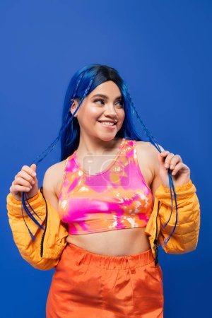 beauty trends, dyed hair, cheerful female model with blue hair posing in puffer jacket on blue background, vibrant color, urban fashion, individualism, young woman smiling and looking away