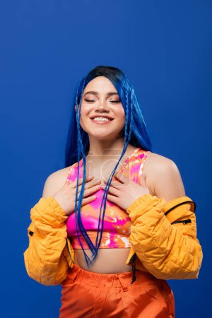 fashion trends, dyed hair, happy female model with blue hair posing in puffer jacket on blue background, vibrant color, urban fashion, individualism, young woman smiling with closed eyes