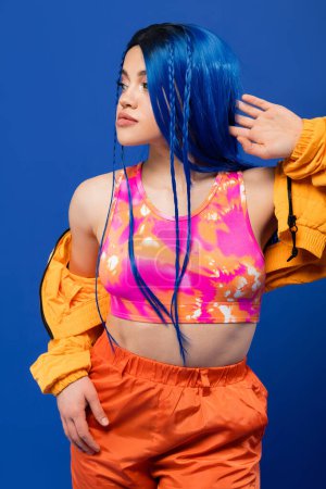bunte Kleidung, gefärbte Haare, weibliches Model mit blauen Haaren posiert in Pufferjacke auf blauem Hintergrund, lebendige Farbe, urbane Mode, Individualismus, junge Frau mit flippigem Look 