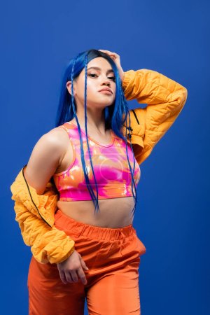 Generation z, gefärbte Haare, weibliches Model mit blauen Haaren posiert in Pufferjacke auf blauem Hintergrund, lebendige Farbe, urbane Mode, Individualismus, junge Frau mit flippigem Look 