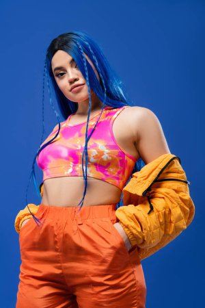 colorful clothes, dyed hair, female model with blue hair posing with hands in pockets on blue background, puffer jacket, vibrant color, urban fashion, individualism, young woman with funky look 