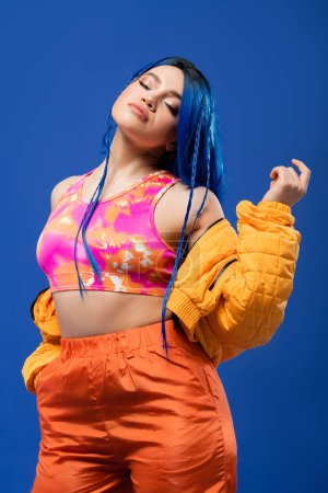 colorful clothes, dyed hair, female model with blue hair posing in puffer jacket on blue background, hand in pocket, vibrant color, urban fashion, individualism, young woman with funky look 