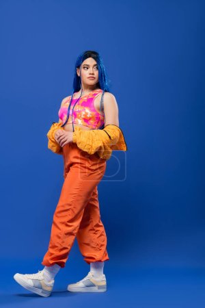 volle Länge, Frau mit gefärbten Haaren, modisches Statement, tätowiertes weibliches Model mit blauen Haaren posiert in Pufferjacke und orangefarbener Hose auf blauem Hintergrund, lebendige Farbe, urbane Mode, Individualismus 