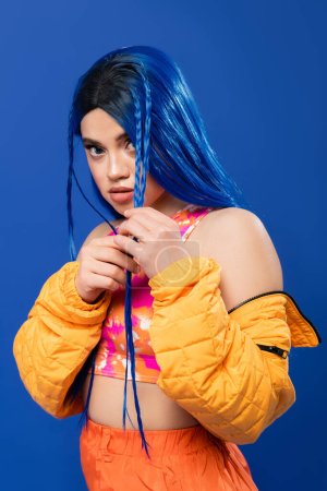 bunte Kleidung, junge Frau mit gefärbten Haaren posiert in Pufferjacke auf blauem Hintergrund, lebendige Farbe, urbane Mode, Individualismus, junge Frau mit rebellischem Stil, weibliches Modell mit blauen Haaren 