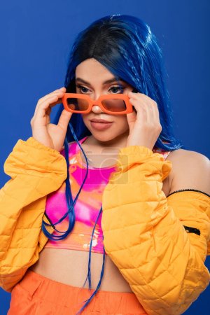mode et style, jeune mannequin femme aux cheveux bleus et tresses portant des lunettes de soleil orange isolées sur fond bleu, génération z, style rebelle, vêtements colorés, individualisme, femme moderne 