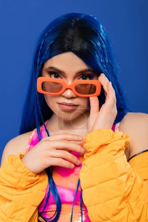 fashion and style, young female model with blue hair and braids wearing trendy sunglasses isolated on blue background, generation z, rebel style, individualism, modern woman looking at camera