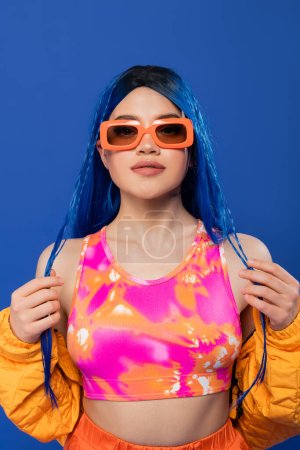 Mode-Statement, junges weibliches Model mit blauen Haaren und Zöpfen posiert mit trendiger Sonnenbrille auf blauem Hintergrund, Generation Z, Rebellentyp, bunte Kleidung, Individualismus, moderne Frau 