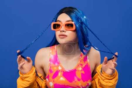 fashion statement, young female model with blue hair touching braids and trendy sunglasses isolated on blue background, generation z, rebel style, colorful clothes, individualism, modern woman 