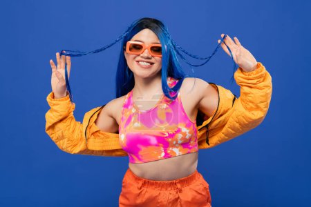 fashion statement, cheerful female model with blue hair and trendy sunglasses isolated on blue background, generation z, rebel style, colorful clothes, individualism, modern positive woman 