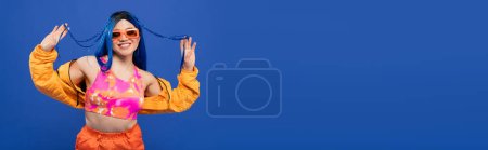 Photo for Fashion statement, positive female model with blue hair and trendy sunglasses isolated on blue background, generation z, rebel style, colorful clothes, individualism, modern woman, banner - Royalty Free Image