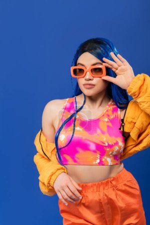 Photo for Fashion statement, young female model with blue hair adjusting trendy sunglasses isolated on blue background, generation z, rebel style, colorful clothes, individualism, modern woman - Royalty Free Image
