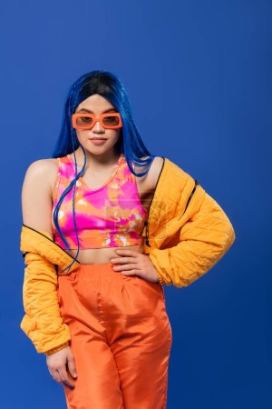 trendiges Accessoire, junges weibliches Model mit blauen Haaren und trendiger Sonnenbrille posiert mit der Hand an der Hüfte isoliert auf blauem Hintergrund, rebellischer Stil, bunte Kleidung, Individualismus, moderne Frau 