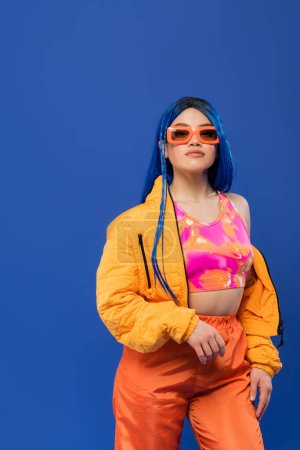 gen z, fashion statement, young female model with blue hair adjusting trendy sunglasses isolated on blue background, rebel style, colorful clothes, individualism, modern woman 