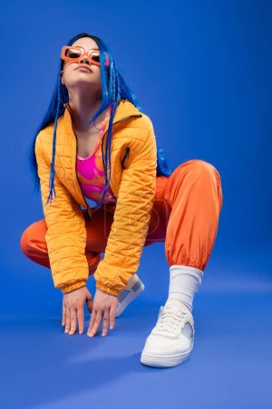 full length of young female model with blue hair and trendy sunglasses sitting on haunches on blue background, generation z, rebel style, individualism, modern fashion, trendy accessory 