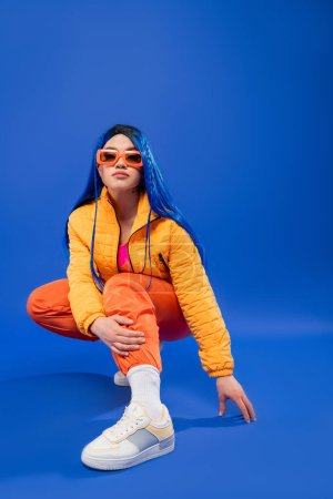 individualism, full length of pretty female model with blue hair and trendy sunglasses sitting on haunches on blue background, rebel style, modern fashion, trendy accessory, generation z, 