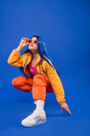 generation z, full length of pretty female model with blue hair and trendy sunglasses sitting on haunches on blue background, rebel style, modern fashion, trendy accessory, gen z