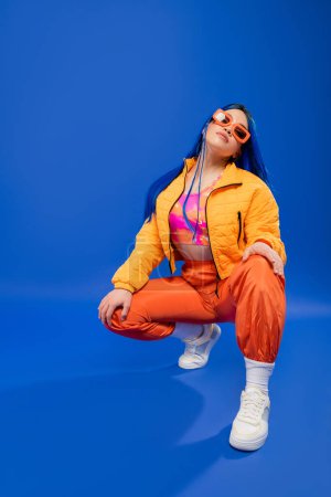 Photo for Fashion statement, full length of pretty female model with blue hair and trendy sunglasses sitting on haunches on blue background, rebel style, modern fashion, trendy accessory, gen z - Royalty Free Image