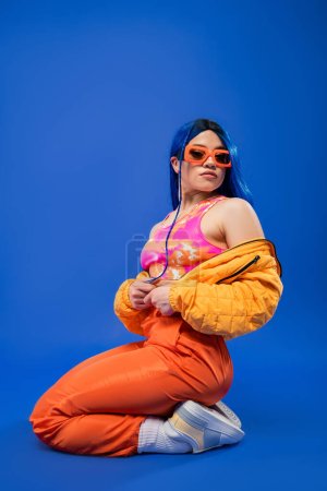 fashion statement, full length of beautiful female model with blue hair and trendy sunglasses sitting on blue background, rebel style, modern fashion, trendy accessory, youth 