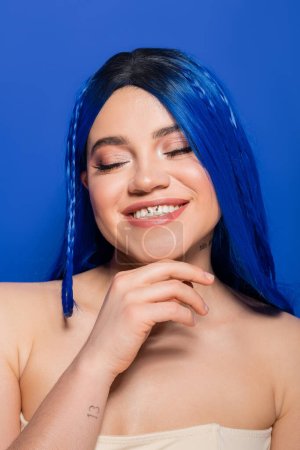 Foto de Concepto de tendencias de belleza, mujer joven feliz con el pelo teñido posando sobre fondo azul, color de pelo, individualismo, modelo femenino con maquillaje y peinado de moda sonriendo con los ojos cerrados, juventud vibrante - Imagen libre de derechos