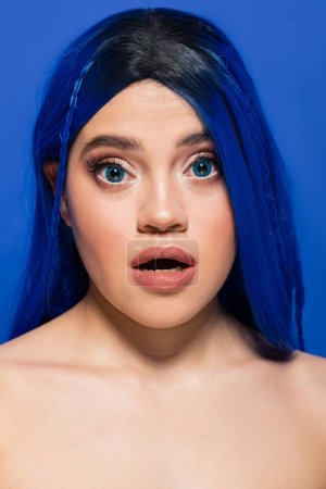 Konzept der Schönheitstrends, Porträt einer schockierten jungen Frau mit gefärbten Haaren, die auf blauem Hintergrund posiert, Haarfarbe, Individualismus, weibliches Model mit Make-up und trendiger Frisur, lebendige Jugend, emotionale 