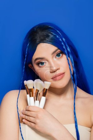 beauty industry, individualism, young woman with vibrant hair and eyes looking at camera while holding cosmetic brushes on blue background, makeup, beauty trends, visage, youth, self expression 