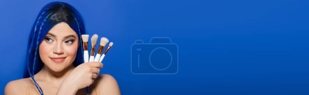 beauty industry, individualism, happy young woman with vibrant hair and eyes looking away while holding cosmetic brushes on blue background, makeup, beauty trends, visage, self expression, banner 