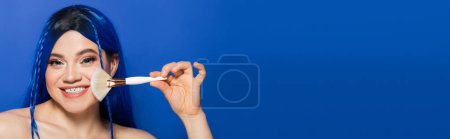 Photo for Beauty trends, individualism, happy young woman with vibrant eyes and hair looking at camera while holding makeup brush on blue background, cosmetic, self expression, visage, youth, banner - Royalty Free Image