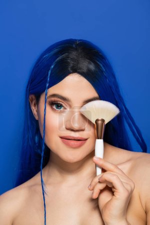 beauty trends, individualism, young woman with vibrant hair looking at camera while holding makeup brush near eye on blue background, cosmetic, self expression, visage, youth