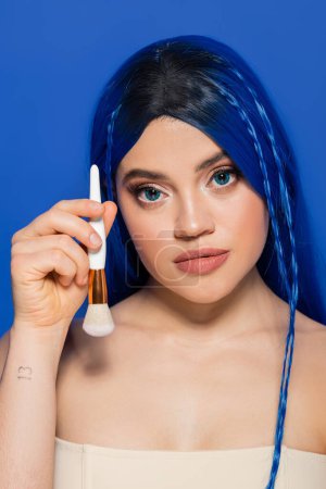Photo for Beauty trends, individualism, young woman with vibrant eyes and hair looking at camera while holding cosmetic brush on blue background, makeup, self expression, visage, youth - Royalty Free Image