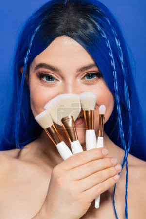 Make-up-Tools, jugendliche Haut, fröhliche junge Frau mit lebendigen Haaren und Augen, die Kosmetikpinsel auf blauem Hintergrund hält, Make-up, Beauty-Trends, Visage, Selbstausdruck, Schönheitsindustrie, Coverface 