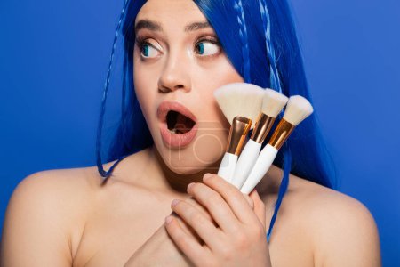Make-up-Tools, jugendliche Haut, schockierte junge Frau mit lebendigen Haaren und Augen, die Kosmetikpinsel auf blauem Hintergrund hält, Make-up, Beauty-Trends, Visage, Selbstausdruck, Schönheitsindustrie 