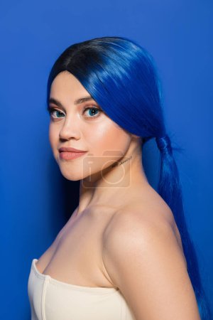 glowing skin concept, portrait of tattooed young woman with vibrant hair color posing with bare shoulders on bright blue background, youth, individualism, beauty trends, unique identity 
