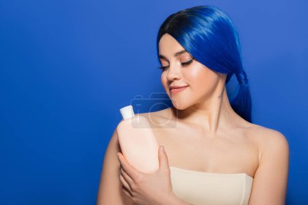 Téléchargez les photos : Concept de soins du corps et des cheveux, portrait de jeune femme tatouée avec une couleur de cheveux vibrante posant avec des épaules nues sur fond bleu, tenant bouteille cosmétique avec shampooing, tendances beauté - en image libre de droit