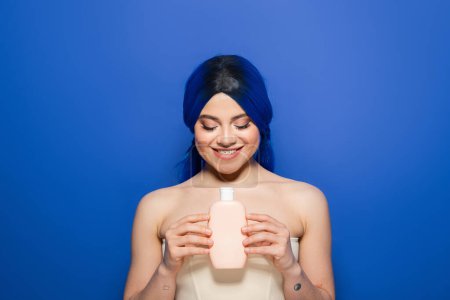 Haarpflegekonzept, Porträt einer fröhlichen jungen Frau mit lebendiger Haarfarbe, die mit nackten Schultern auf blauem Hintergrund posiert, Kosmetikflasche mit Shampoo in der Hand, Beauty-Trends, gesundes Haar 