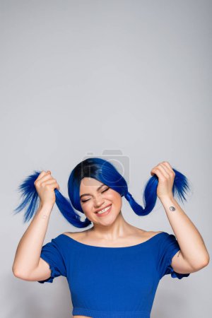 Foto de Positividad y energía juvenil, mujer tatuada y feliz con ojos cerrados y cabello teñido sonriendo sobre fondo gris, peinado, pelo azul, belleza moderna, expresión personal, individualismo - Imagen libre de derechos