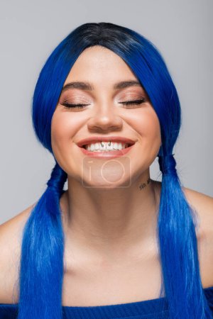 positivity and youth, tattooed woman with closed eyes and blue hair smiling on grey background, hairstyle, vibrant color, modern beauty, self expression, individualism 