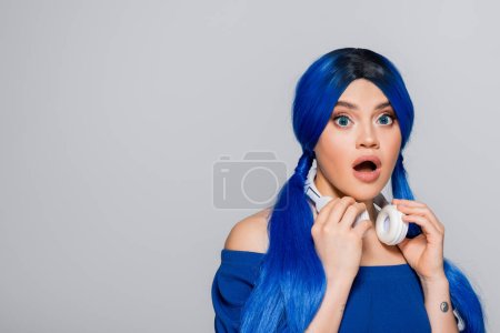 Foto de Amante de la música, mujer joven sorprendida con el pelo azul y auriculares inalámbricos sonriendo sobre fondo gris, juventud vibrante, individualismo, subcultura moderna, expresión personal, tatuaje, sonido - Imagen libre de derechos
