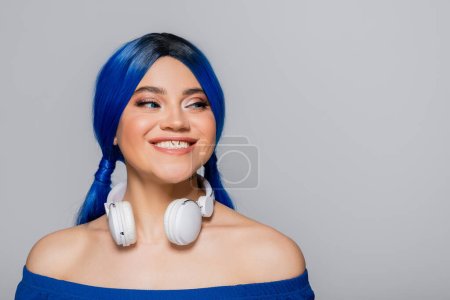 Musikliebhaberin, lächelnde junge Frau mit blauen Haaren und kabellosen Kopfhörern, die auf grauem Hintergrund lächelt, lebendige Jugend, Individualismus, moderne Subkultur, Selbstausdruck, Tätowierung, Sound 