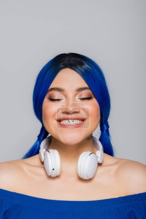 Musikliebhaberin, positive junge Frau mit blauen Haaren und kabellosen Kopfhörern, die auf grauem Hintergrund lächelt, lebendige Jugend, Individualismus, moderne Subkultur, Selbstausdruck, Tätowierung, Sound 