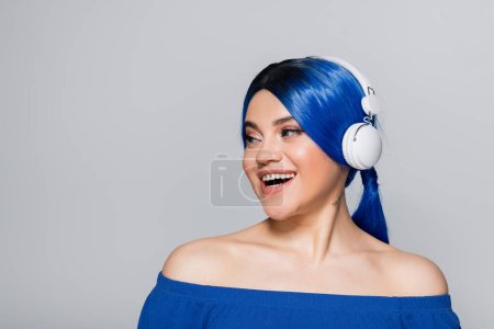 expresión personal, amante de la música, joven feliz con cabello azul escuchando música en auriculares inalámbricos sobre fondo gris, juventud vibrante, individualismo, subcultura moderna, tatuaje, sonido 