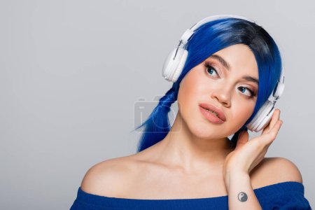 Foto de Expresión personal, mujer joven con pelo azul escuchando música en auriculares inalámbricos sobre fondo gris, juventud vibrante, individualismo, subcultura moderna, tatuaje, sonido, pensativo - Imagen libre de derechos