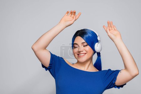 Selbstausdruck, fröhliche junge Frau mit blauen Haaren, die Musik in drahtlosen Kopfhörern auf grauem Hintergrund hört, Tanzen, lebendige Jugend, Individualismus, moderne Subkultur, Tätowierung, Sound 
