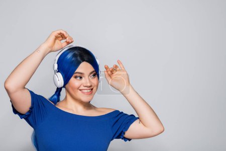 self expression, positive young woman with blue hair listening music in wireless headphones on grey background, vibrant youth, individualism, modern subculture, tattoo, sound 