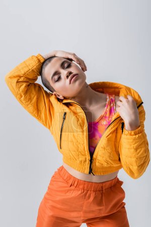 outerwear, individualism, fashion model posing with closed eyes, young woman with short hair standing in yellow puffer jacket on grey background, isolated, youth culture, casual wear