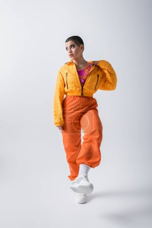 stylish look, outerwear, casual attire, fashion model posing in yellow puffer jacket and bright pants on grey background, woman with short hair running and looking away, modern subculture 