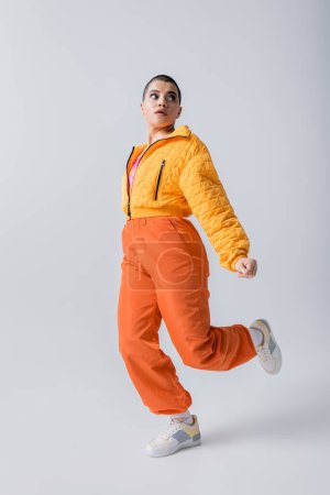 stylisches Aussehen, Oberbekleidung, Freizeitkleidung, Modemodel posiert in gelber Pufferjacke und orangefarbener Hose auf grauem Hintergrund, Frau mit kurzen Haaren läuft und schaut weg, moderne Subkultur 