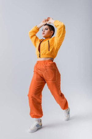 modern subculture, outerwear, casual attire, fashion model posing in yellow puffer jacket and orange pants on grey background, woman with short hair with raised hands, stylish look, individualism 