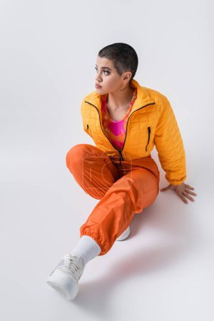 fashion statement, outerwear, female model posing in casual attire, young woman with short hair and puffer jacket sitting on grey background, looking away, personal style, self expression 